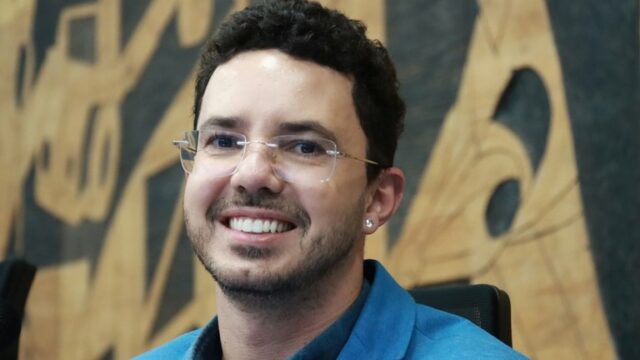 Homem sorrindo, com óculos sem armação e paletó azul, em sessão na Câmara Municipal de Ponta Grossa