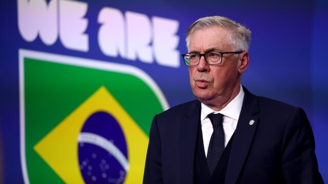 O técnico Carlo Ancelotti veste terno escuro e óculos em coletiva de imprensa com o escudo da seleção ao fundo
