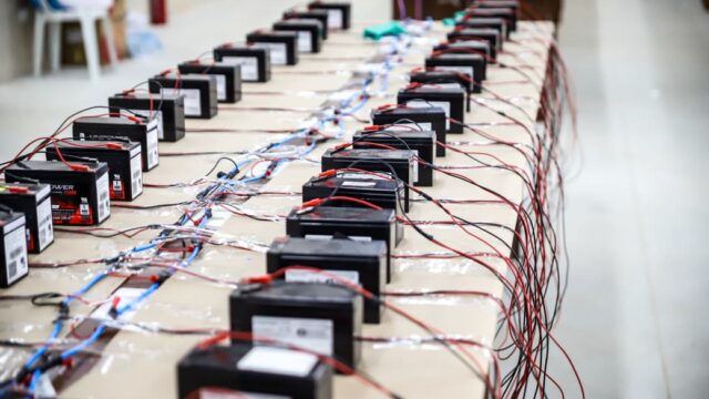 Fileira de baterias pretas conectadas a fios vermelhos e pretos sobre mesas para recarga