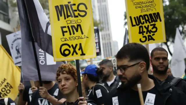 Pessoas seguram cartazes amarelos com inscrições sobre a jornada de trabalho durante manifestação em rua