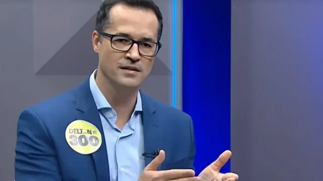 Deltan Dallagnol veste terno azul e camisa clara, gesticulando em estúdio com fundo neutro