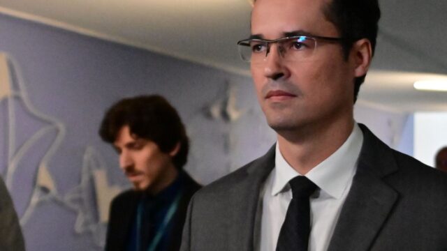 Deltan Dallagnol usando terno cinza, camisa branca e gravata preta em ambiente interno