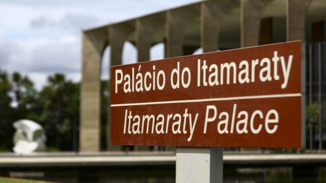 Placa marrom com a inscrição Palácio do Itamaraty e Itamaraty Palace em destaque com a fachada do prédio ao fundo