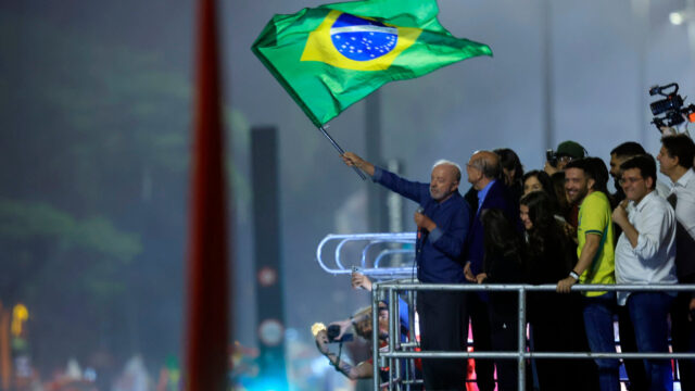 Lula acenando e segurando uma bandeira do Brasil em cima de um palanque cercado por apoiadores