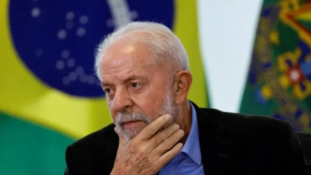 Primeiro plano do presidente Luiz Inácio Lula da Silva com a mão no queixo e bandeira do Brasil ao fundo