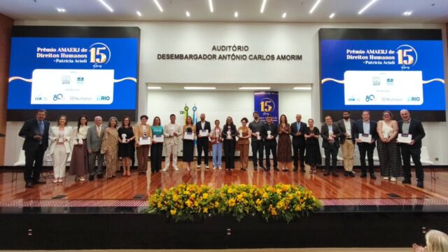 Grupo de premiados com troféus e certificados no palco de auditório com telões azuis ao fundo