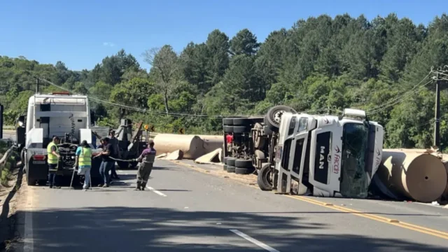 Caminhão branco da marca MAN tombado na lateral da rodovia com bobinas de papel e guincho prestando atendimento