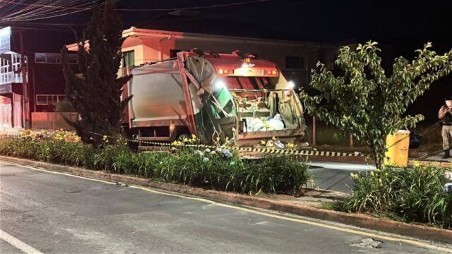 Caminhão de coleta de lixo parado à noite em rua com vegetação e fita de isolamento de trânsito