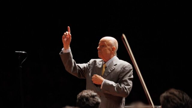 Maestro careca de terno cinza regendo com o braço levantado em um concerto musical