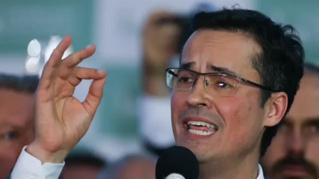 Deltan Dallagnol de óculos fala ao microfone com a mão direita levantada em gesto